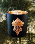 Saint O'Niell Alchemy Forest Bathing luxury scented candle in black matte vessel with gold metallic interior bearing a decorative scrolled fleur de lys label and surrounded by dark forest trees to convey the full-bodied, dark, authentic Mossy Woods scent profile of this aristocratic candle.