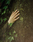 A hand reaching out from the ivy and resting on a moss-covered tree trunk to convey Saint O'Niell Alchemy Forest Bathing luxury scented candle with a Mossy Woods olfactory scent pyramid.