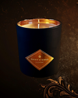 Saint O'Niell Alchemy Grand Cru Chocolat luxury scented candle in black matte vessel with metallic gold interior bearing the original gold foil diamond shaped label. The exquisitely crafted artisan candle is set against a richly textured brown background with decorative gold scrolling on the right side and bottom.