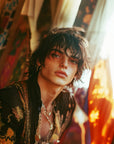 Saint O'Niell Alchemy Haute Bohème luxury scented candle created for "The Troubadour" as depicted by this beautiful artistic, rock star, young man with toussled hair, earrings and neclaces wearing a silk velvet robe against a backdrop of warm, mutil-colored draperies.