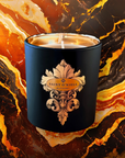 Saint O'Niell Alchemy Haute Bohème luxury scented candle in black matte vessel with gold metallic interior bearing decorative scrolled fleur de lys label with gold foil logo treatment against a warm, resinous backdrop of marble in varying shades of orange, brown, ivory and black to convey the embered spices olfactory notes pyramid it is exquisitely crafted with.