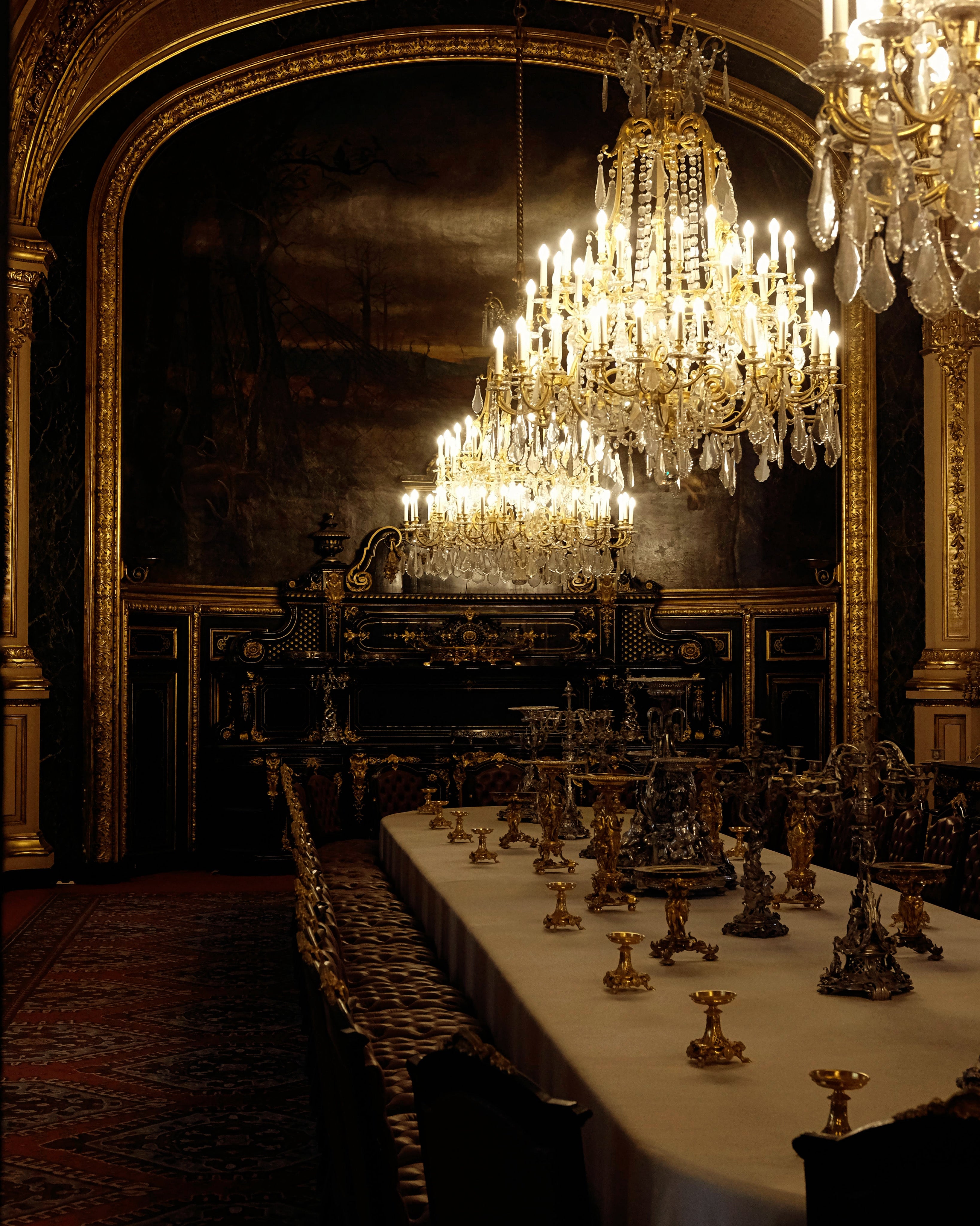 Dark, gilded dining room in historic mansion let by crystal chandeliers conveying elegance of Saint O'Niell's Manor Born luxury scented candle of waxed woods, limoncello and moss.