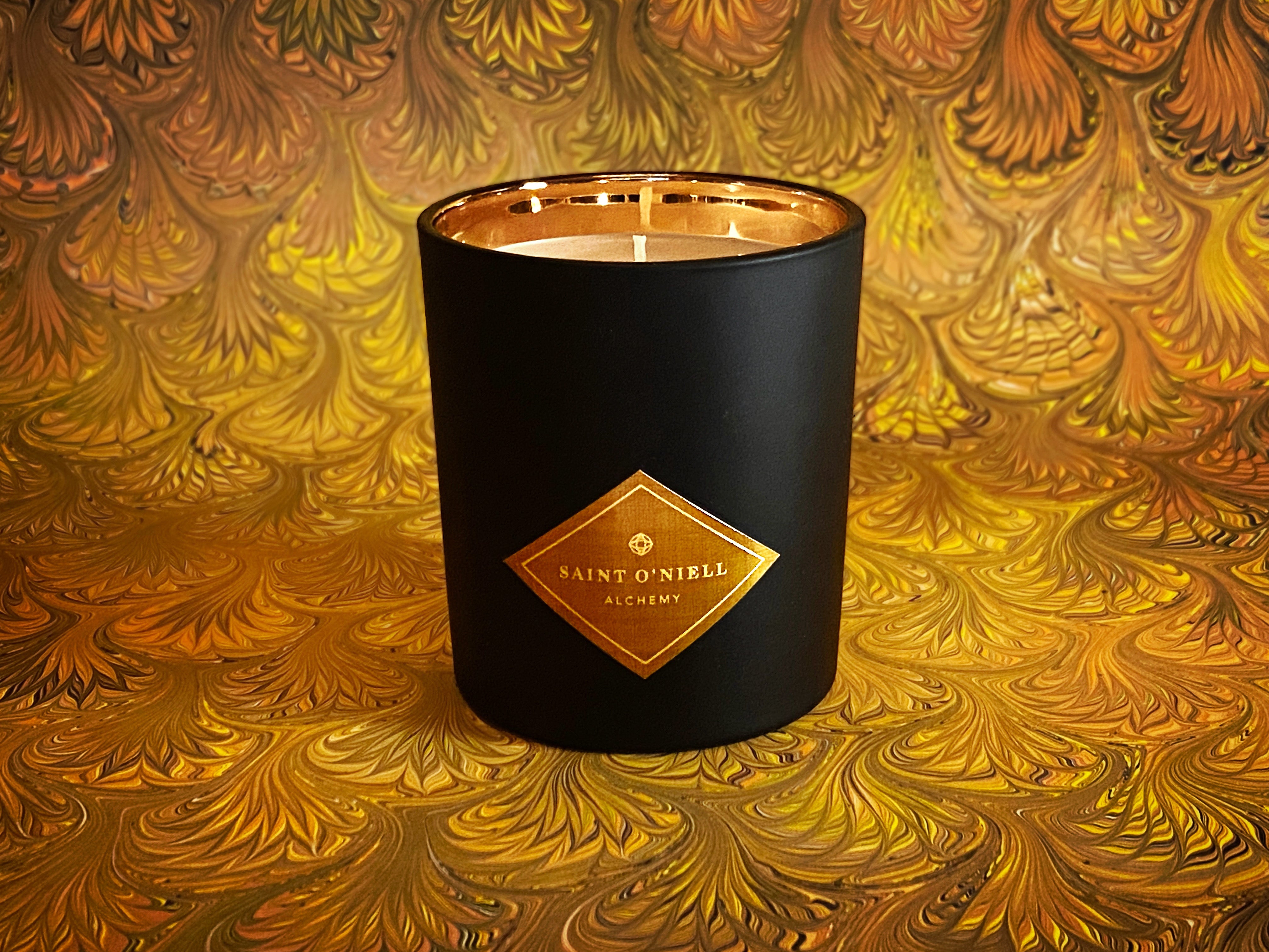 Saint O'Niell Alchemy's classic luxury scented candle in black matte vessel with gold metallic interior on a backdrop of antiqued Italian marbled paper in shades of yellow, gold, brown and orange conveys the woody chypre notes of the best selling Manor Born scented candle.
