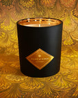 Saint O'Niell Alchemy's classic luxury scented candle in black matte vessel with gold metallic interior on a backdrop of antiqued Italian marbled paper in shades of yellow, gold, brown and orange conveys the woody chypre notes of the best selling Manor Born scented candle.