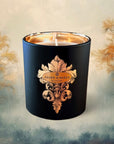 Saint O'Niell Alchemy Sylvan luxury scented candle in black matte vessel with gold metallic interior bearing the decorative fleur de lys scroll label with gold foil oval containing logo set against a painting of foggy forest with golden birch peaking out at both sides and green bushes at bottom.