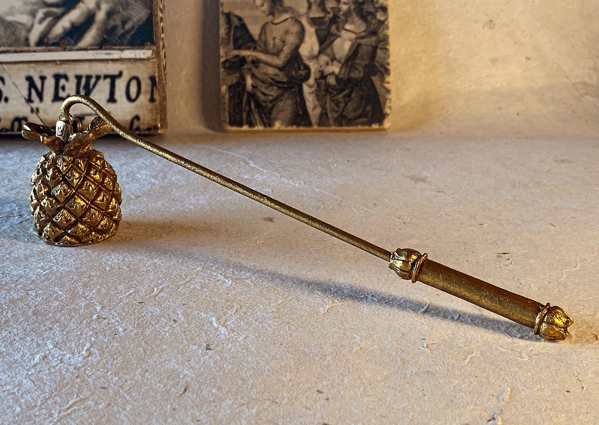 Rearview of the Gold Pineapple Candle Snuffer showing the intricately forged handle atop a natural surface with a backdrop of Renaissance art to convey the natural charm and elegance of the Saint O'Niell Alchemy brand.