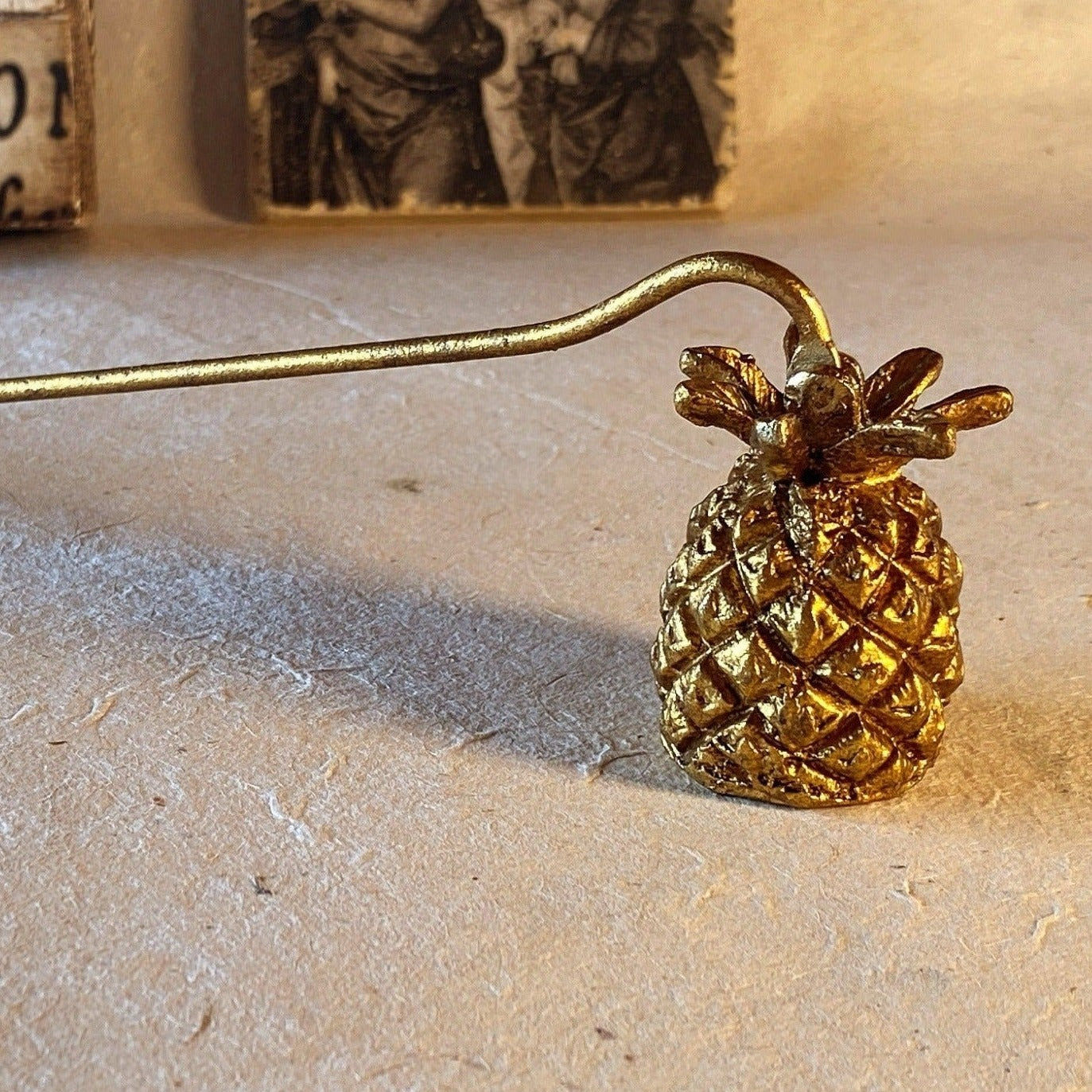 The hand-finished metal Pineapple Candle Snuffer in antique gold sits atop a natural surface with a backdrop of Renaissance art to convey the natural charm and classic style of the Saint O'Niell Alchemy brand.