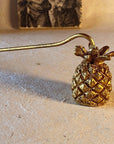 The hand-finished metal Pineapple Candle Snuffer in antique gold sits atop a natural surface with a backdrop of Renaissance art to convey the natural charm and classic style of the Saint O'Niell Alchemy brand.