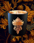 Saint O'Niell Alchemy Manor Born luxury scented candle in black matte vessel with metallic gold interior bearing a decorative fleur de lys scoll label with gold foil oval containing logo against a artistic backdrop of plants painted in gold, green, purple and black to convey its wood chypre olfactory profile.