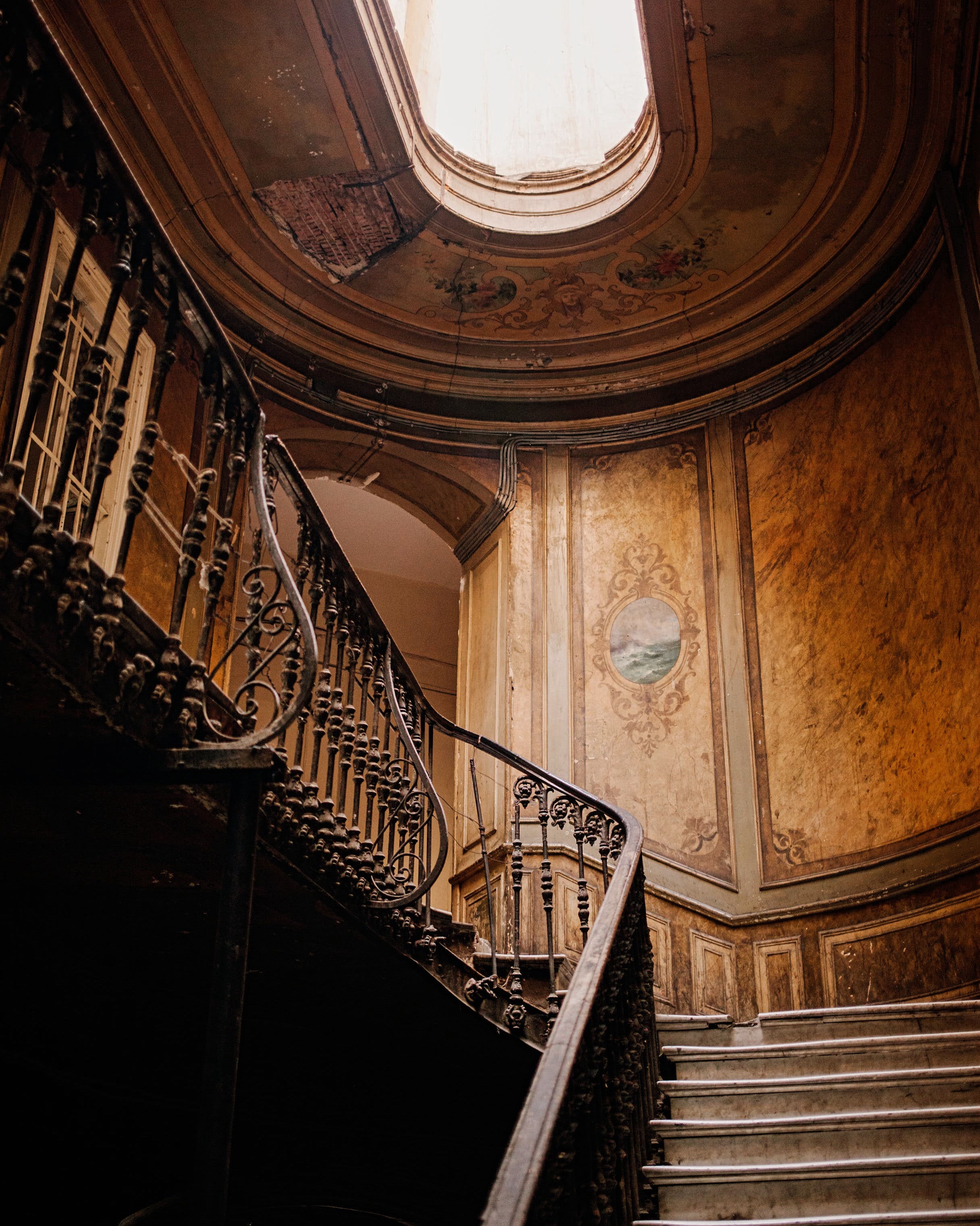 A burlwood paneled wall with inset mural along curved staircase with wrought-iron bannister and skylight inside historic mansion conveys the woody chypre notes of the elegant Manor Born scented candle by Saint O'Niell Alchemy.