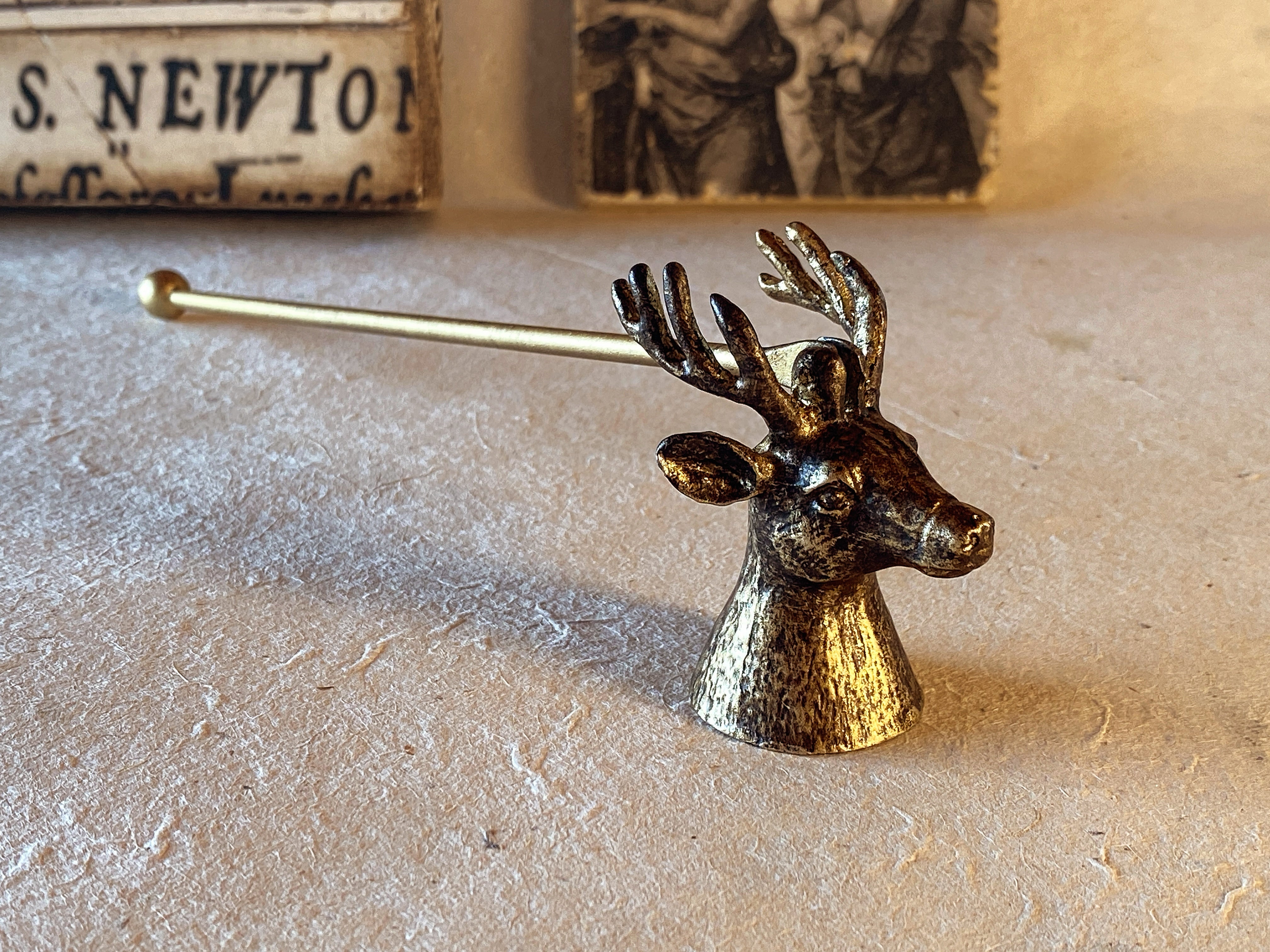 The hand-finished metal Stag's Head Candle Snuffer in an antique gold finish sits atop a natural surface with a backdrop of Renaissance art to convey its natural charm and classic style.